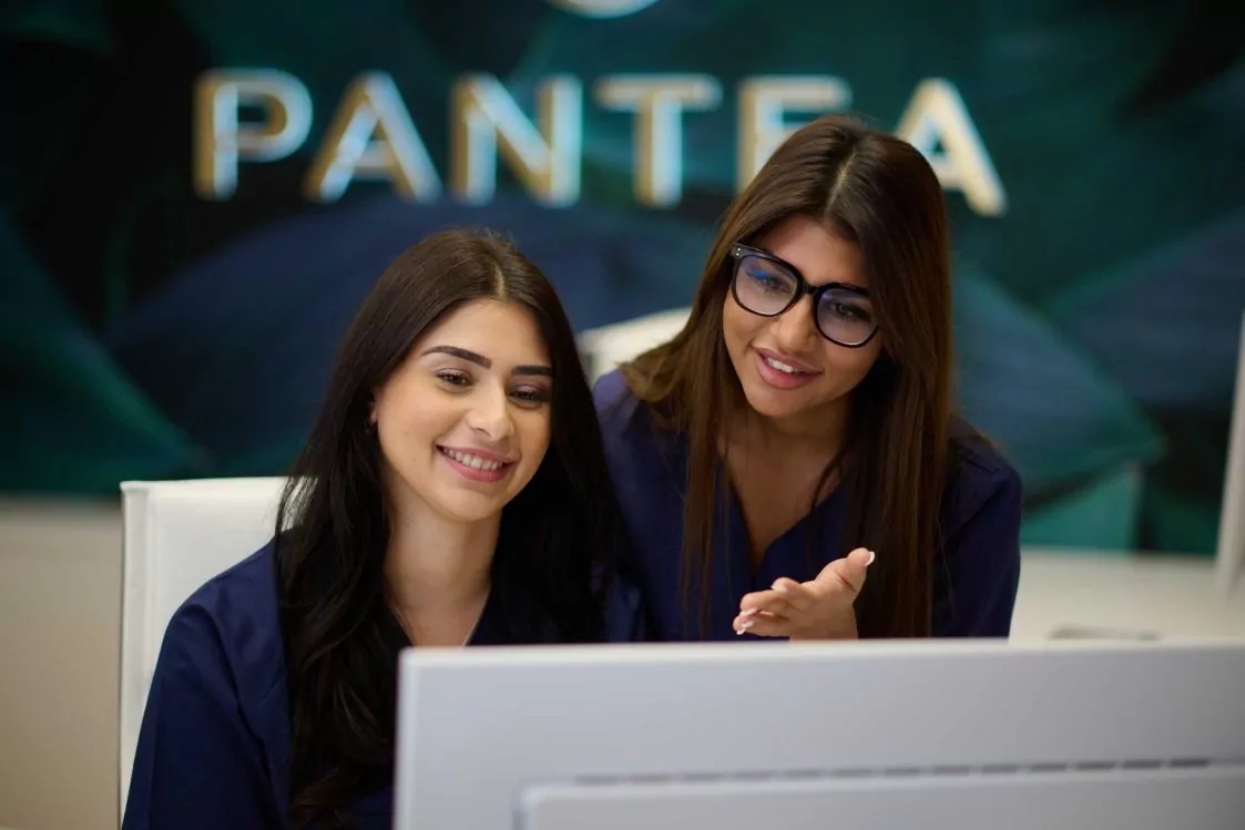 Zwei Frauen in blauen Blusen sitzen vor einem Computer, im Hintergrund ist ein Schild mit der Aufschrift 'PANTEA'.